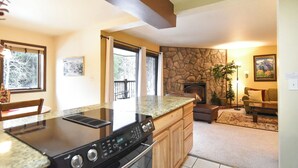 Una cocina moderna con encimera de granito, horno empotrado y armarios de madera. Cuenta con una chimenea de piedra, un comedor con mesa y sillas, y una sala de estar con sofá y alfombra.