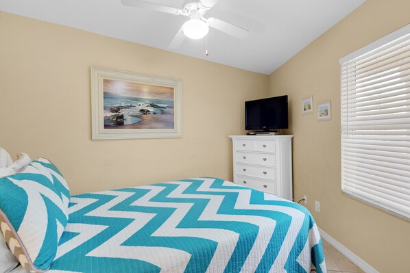 A bedroom with a bed, a ceiling fan, a dresser, a television, and a window with blinds.