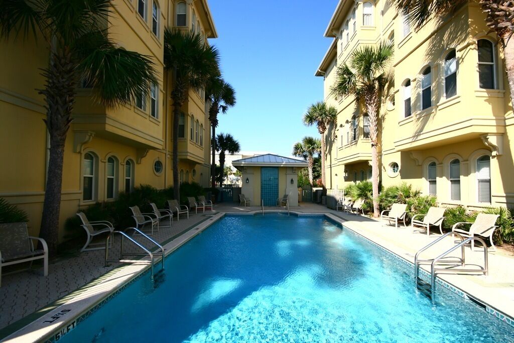 A swimming pool surrounded by yellow buildings with palm trees, lounge chairs, and a clear blue sky.