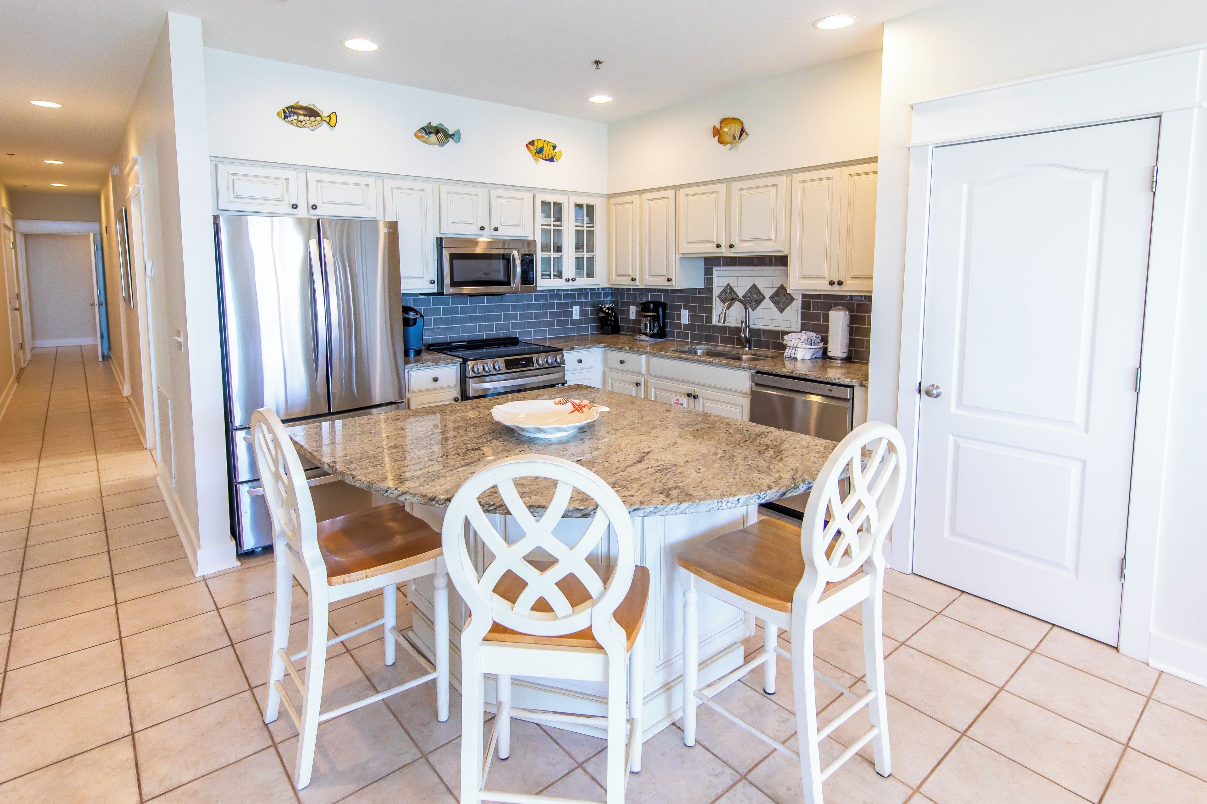A kitchen with white cabinets, a stainless steel refrigerator, a microwave, and a stove. There are two white chairs with wooden seats and backs.