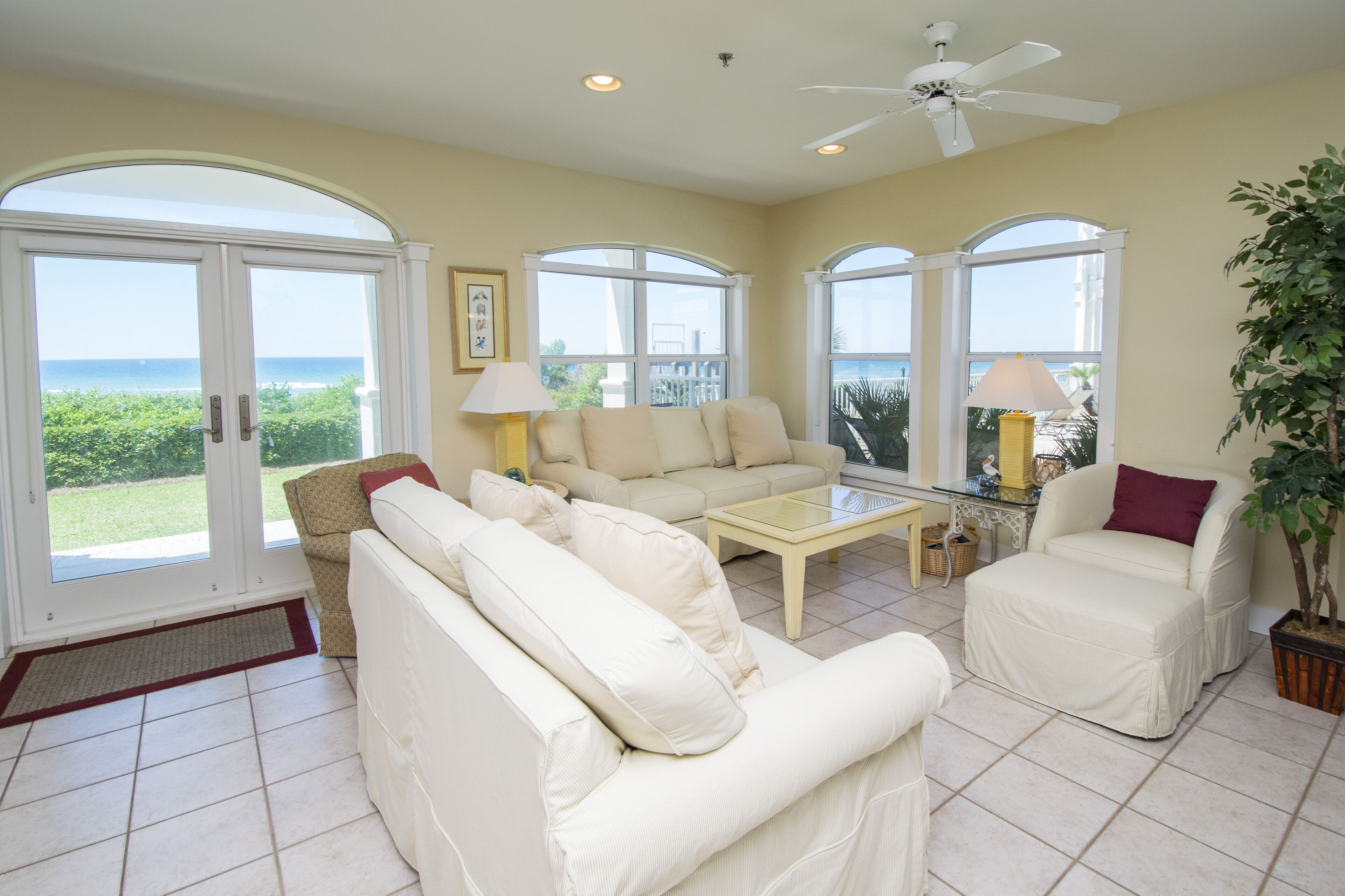 A spacious living room with a large window overlooking the ocean, a ceiling fan, and a potted plant.