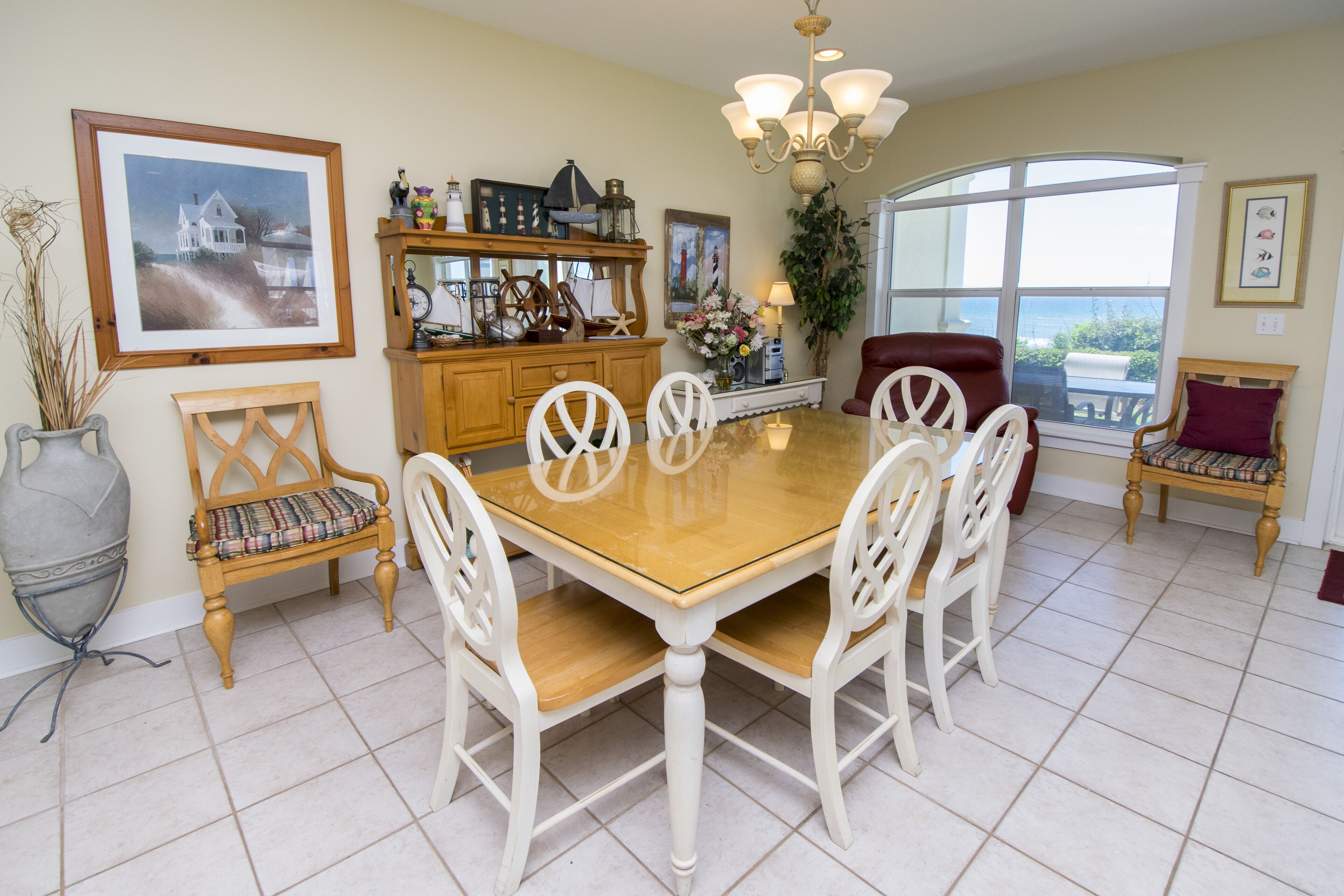 Uma sala de jantar com mesa de madeira e cadeiras brancas, um candelabro e uma janela grande com vista.