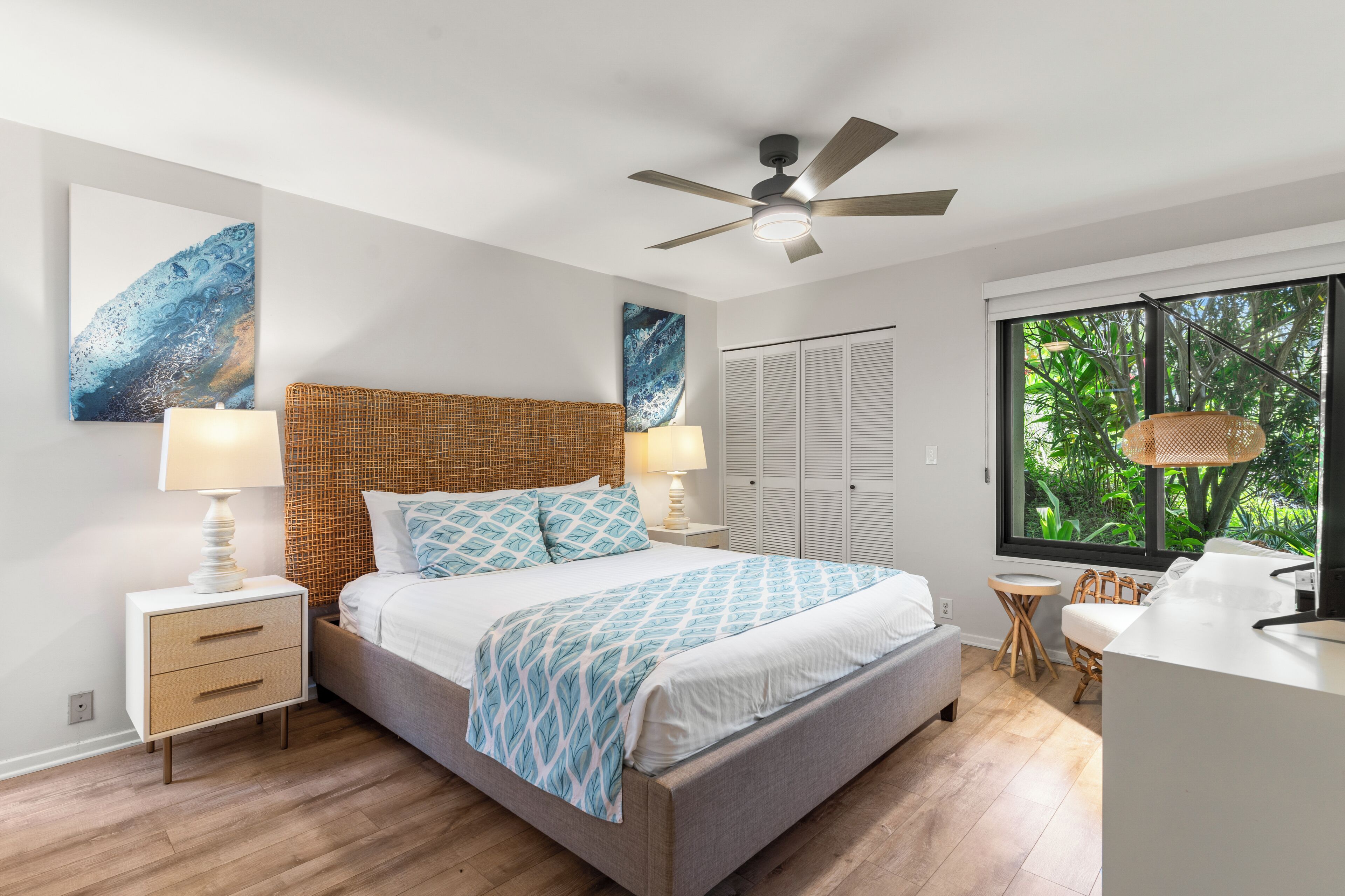 A modern bedroom with a bed, nightstands, a ceiling fan, and a view of greenery through the window.