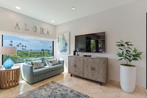 A living room with a sofa, a cabinet, a large window with a view, a TV, and a plant. - Exquisite Escape: Elegantly Renovated Wailea Elua 801 – Tranquil Paradise (Wailea, Maui)