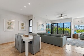A modern living room with a dining area, a gray sofa, and a large window offering a view of palm trees. - Exquisite Escape: Elegantly Renovated Wailea Elua 801 – Tranquil Paradise (Wailea, Maui)