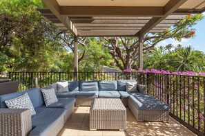 A covered outdoor seating area with a sectional sofa and ottomans, surrounded by a metal railing and lush greenery.