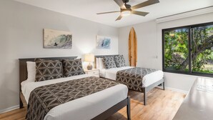 A bedroom with two beds, a ceiling fan, artwork, and a window with a view of greenery.