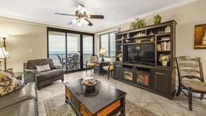 A living room with a sofa, coffee table, TV stand, and a view of the ocean through a sliding glass door. - Broadmoor 105 by Liquid Life (Orange Beach)