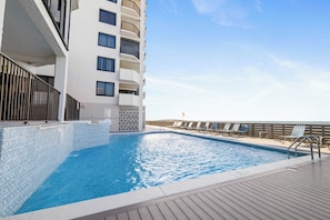 A swimming pool with a wooden deck, a white building with balconies, and a clear sky.
