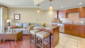 A modern living room with a sofa, coffee table, and ottoman, a kitchen with wooden cabinets and a tiled backsplash, and pendant lighting. - Maui Resort Rentals: Honua Kai Konea 515 - Upgraded 5th Floor Oceanview Studio (Lahaina)