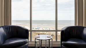 Een kamer met een groot raam met uitzicht op een strand, twee leren fauteuils en een kleine ronde tafel.