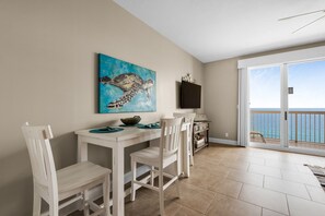 A dining area with a table and chairs, a painting, a television, and a view of the ocean through a sliding glass door. - AMAZING BEACH VIEWS FROM THE SPACIOUS GULF FRONT BALCONY! BEACH CHAIR SERVICE INCLUDED! SUPER CLEAN! WALK TO PIER PARK! (Panama City Beach)