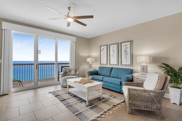 A spacious living room with a large sliding glass door leading to a balcony with a view of the ocean. There is a blue sofa, a white coffee table, and a wicker chair. - AMAZING BEACH VIEWS FROM THE SPACIOUS GULF FRONT BALCONY! BEACH CHAIR SERVICE INCLUDED! SUPER CLEAN! WALK TO PIER PARK! (Panama City Beach)