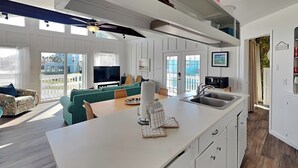 Una cocina moderna con una isla grande, muebles blancos y una zona de comedor con un sofá color turquesa y sillas de mimbre.