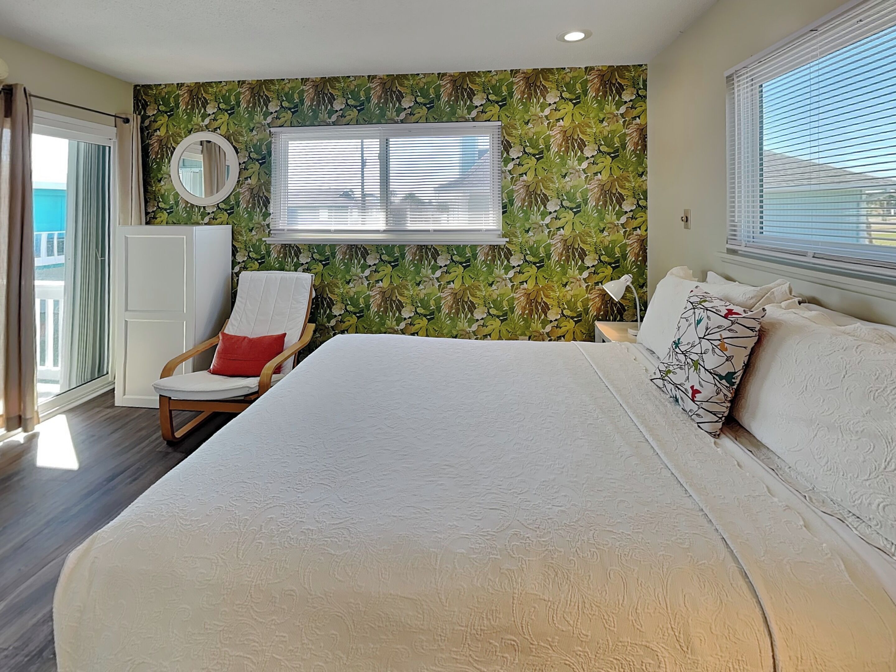 Un dormitorio con una cama grande, una silla con cojín rojo, un espejo en la pared y una ventana con persianas.