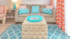 A living room with a sofa, ottoman, and a small side table with a lamp. - Beach Racquet A126 | Steps to the Beach + Pool! (Tybee Island)