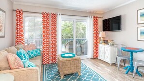 A living room with a sofa, ottoman, and a small table. There is a sliding glass door with curtains, a flat-screen TV mounted on the wall, and a small table with chairs. - Beach Racquet A126 | Steps to the Beach + Pool! (Tybee Island)
