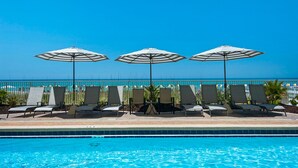 A poolside area with lounge chairs and umbrellas, a clear blue sky, and a view of the ocean.