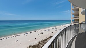 A beach view from a balcony with a clear sky, a sandy beach, and a building with multiple balconies.