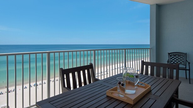 A balcony with a wooden table, two chairs, and a tray with a glass and a bottle. The balcony overlooks a beach with clear blue water and a sandy shore.