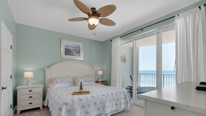 A bedroom with a bed, bedside tables, a ceiling fan, a window with curtains, and a view of the sea.