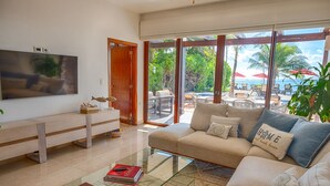 Ein geräumiges Wohnzimmer mit großem Fernseher, bequemen Sitzgelegenheiten und Blick auf Pool und Strand.