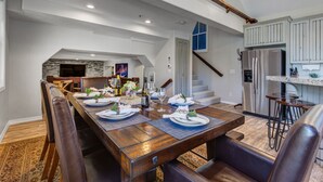 A dining area with a long wooden table set for a meal, a kitchen with modern appliances, and a staircase leading to an upper level. - Epic Home Just Steps to Main Street + Hot tub (Park City)