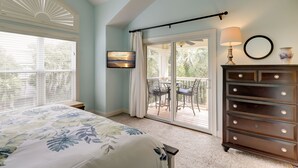 Ein Schlafzimmer mit einem Bett, einer Kommode, einem Deckenventilator, einem Fenster mit Jalousien, einer Schiebetür zu einem Balkon und einem an der Wand montierten Fernseher.