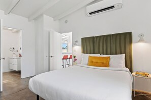Un dormitorio moderno con una cama grande, un equipo de aire acondicionado, una mesita de noche con libros y vistas al exterior.