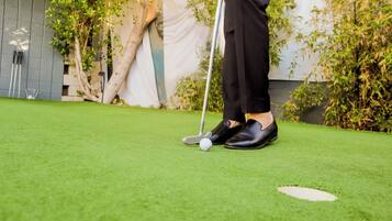 Een persoon staat op kunstgras met een golfbal en club, nabij een witte golfkuil.