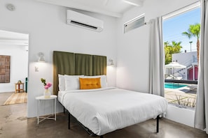 A hotel room with a bed, a small side table, a vase with flowers, a view of a swimming pool, and an air conditioning unit.