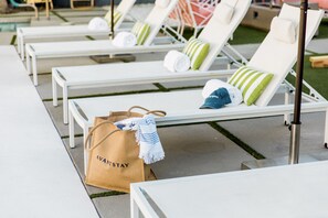 Une rangée de chaises longues blanches, agrémentées de coussins à rayures vertes et blanches, un sac marron portant l’inscription « AVANTSTAY » posé sur le sol, et un poteau métallique au premier plan.