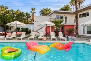 Una piscina con sillones de descanso y sombrillas, rodeada por un edificio con balcón.