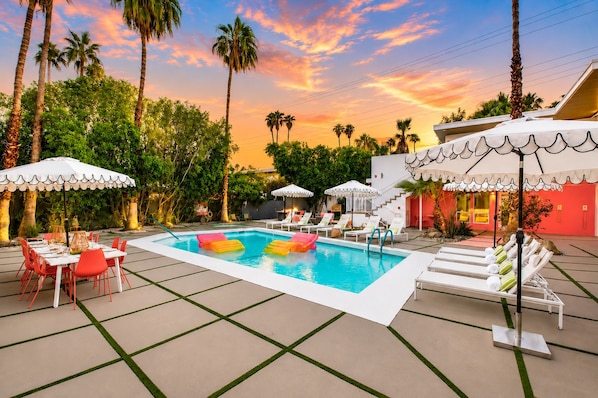 A swimming pool with lounge chairs and umbrellas, surrounded by palm trees and a sunset sky.