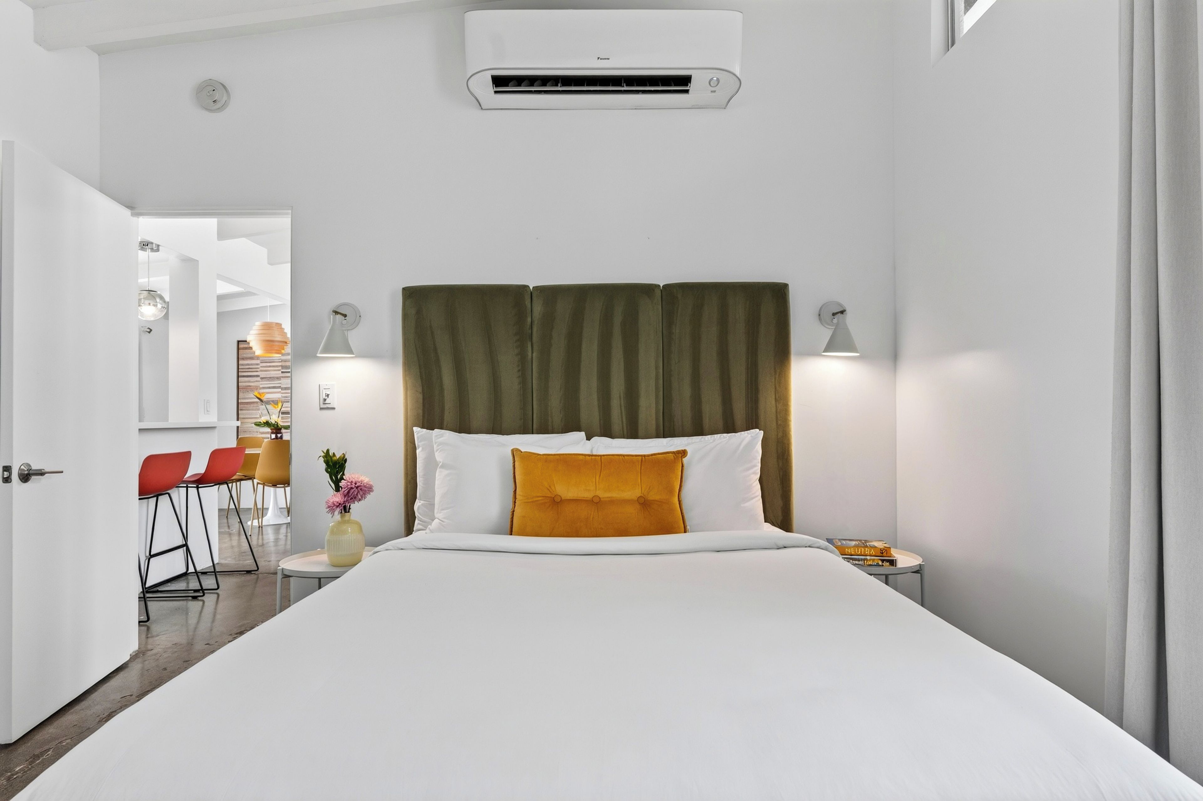 Habitación de hotel con cama, lámparas de noche, unidad de aire acondicionado, una mesita con un libro y vistas al pasillo.