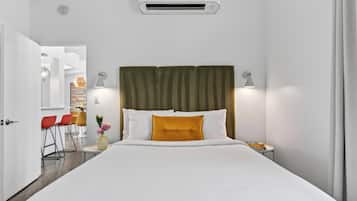 Habitación de hotel con cama, lámparas de noche, unidad de aire acondicionado, una mesita con un libro y vistas al pasillo.