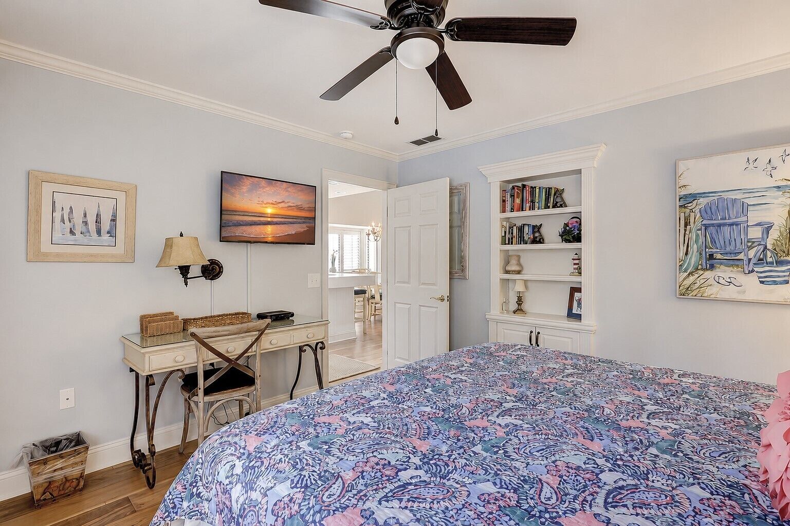 Un dormitorio con cama, escritorio, silla, ventilador de techo, estantería y obras de arte.