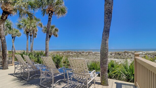 Une terrasse avec des chaises longues donnant sur une plage bordée de palmiers, sous un ciel dégagé.