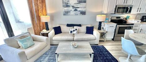 Ein modernes Wohnzimmer mit einer Couch, einem Couchtisch und einem Essbereich, verbunden mit einer Küche mit weißen Schränken und Geräten.