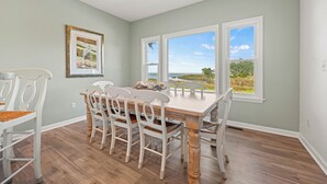 Un área de comedor con una mesa y sillas, ventanas amplias con vistas al exterior y un cuadro enmarcado en la pared.
