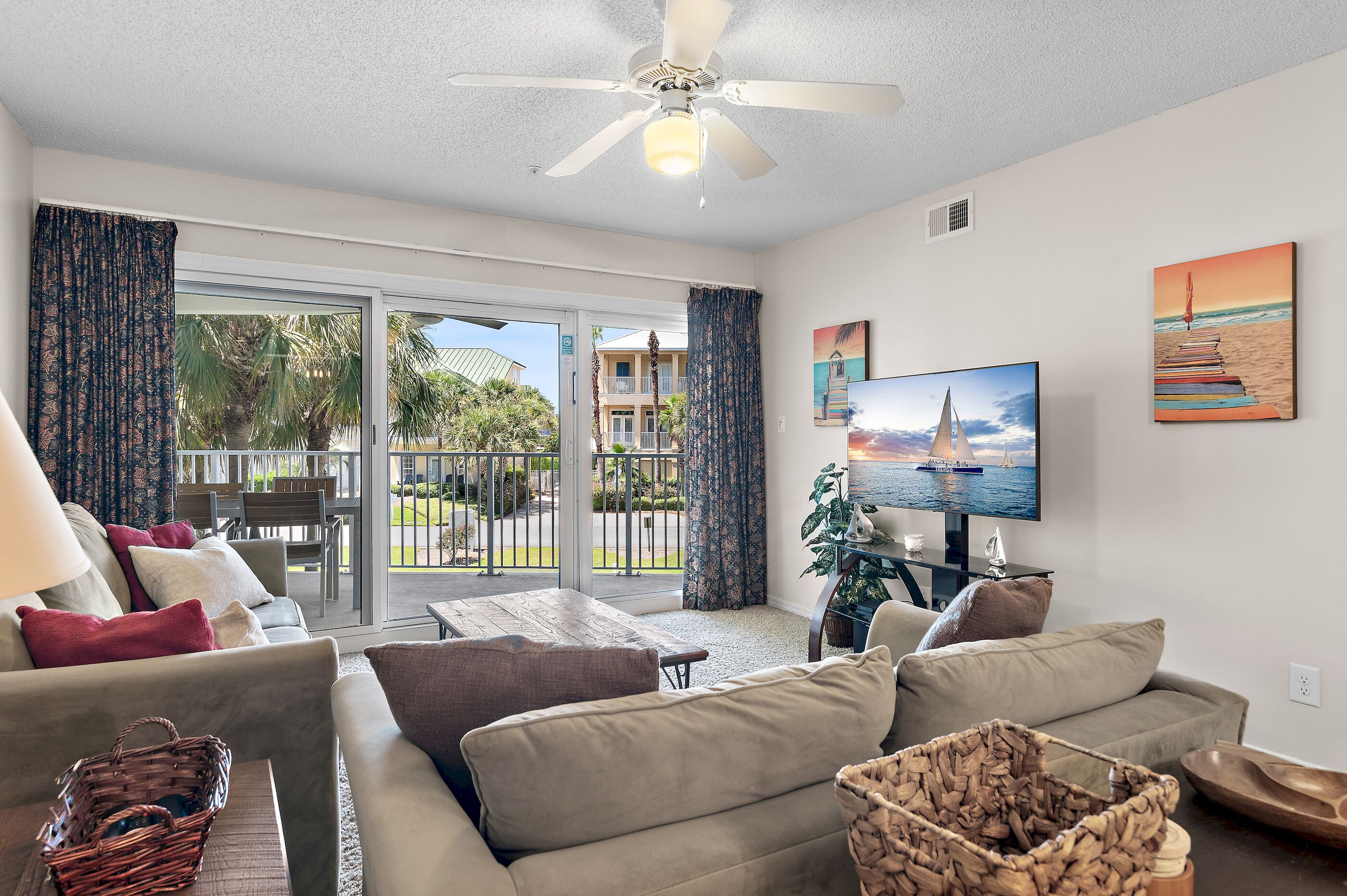A living room with a sofa, a TV, and a balcony with a view of palm trees and a building.