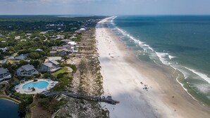 An aerial view of a coastal area with a long sandy beach, a swimming pool, and residential buildings along the shore.