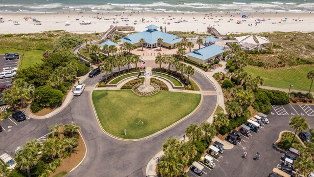 An aerial view of a coastal resort with a central building surrounded by a landscaped area, a parking lot, and a beach in the background.