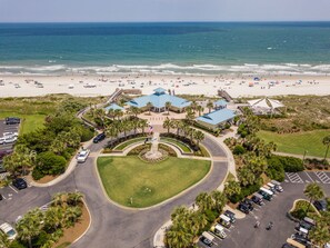 Vista aérea de un resort costero con un edificio central rodeado de jardines, un estacionamiento y una playa al fondo.