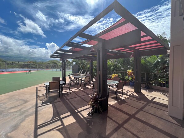 A covered outdoor seating area with tables and chairs, a tennis court in the background, and a view of the surrounding landscape.