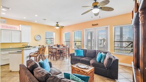 A spacious living area with a brown leather couch, a coffee table, and a dining area with wooden chairs and a table. There are ceiling fans, a clock on the wall, and large windows with blinds. - Villa Laguna | Pools + Boardwalk + Pet Friendly (Port Aransas)