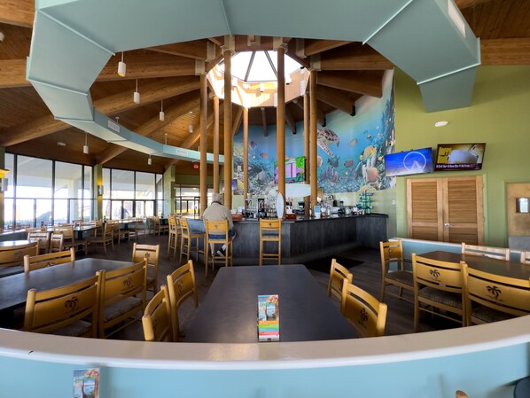 Un restaurante con barra central, mesas y sillas de madera, y un mural de paisajes submarinos en las paredes.