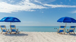 Ein Strand mit zwei blauen Sonnenschirmen und vier Liegestühlen.