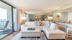 Ein geräumiges Wohnzimmer mit einem großen weißen Sofa, dazu passendem Hocker sowie einem Essbereich mit kleinem Tisch und Stühlen. Ein großes Fenster bietet Blick auf den Ozean.