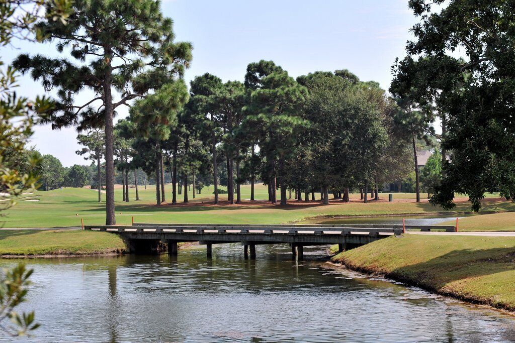 Eine Brücke über ruhiges Wasser, im Hintergrund ein Golfplatz.